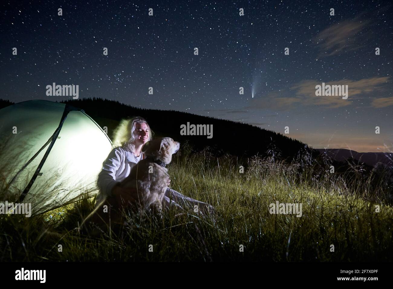 Junge Frau in den Bergen, die eine schöne Sternennacht und einen Kometen beobachtet, sitzt mit ihrem Hund neben dem beleuchteten Zelt auf dem Campingplatz. Speicherplatz kopieren. Konzept der Astrofotografie Stockfoto