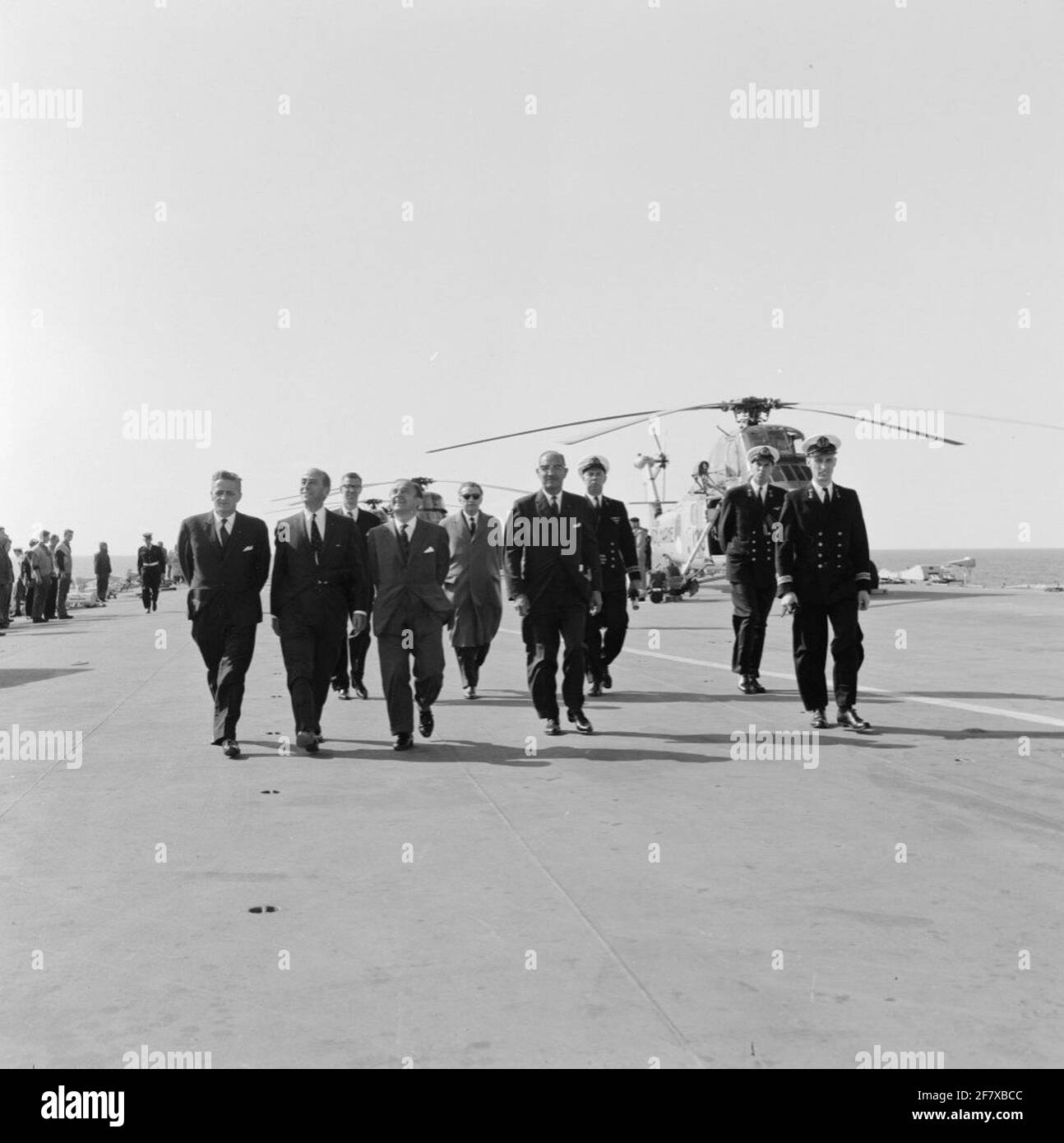 Am 19. September wird das Marijnen-Kabinett (1963-1965) mit dem Flugzeuglager hr.ms segeln. Karel Doorman (R 81) Van Den Helder nach Rotterdam. Einige der Gäste laufen mit einigen Marineoffizieren über die Tür des Türstehers mit einem Transport-/Untersuchungs- und Rettungshubschrauber vom Typ Sikorsky UH-34J im Hintergrund. Der Marine Officer Midden-Behind ist Lieutenant-Sea Kite der 1. Klasse (LTZV 1) van den Kroander. Stockfoto