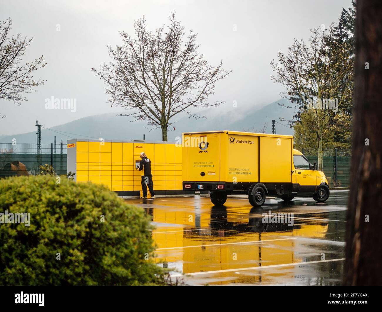 Lahnstein, Deutschland, 10. April 2021: Ein Mitarbeiter der 'Deutsche Post DHL Group' liefert Paket und Pakete mit einem Elektrofahrzeug an eine Packstation Stockfoto