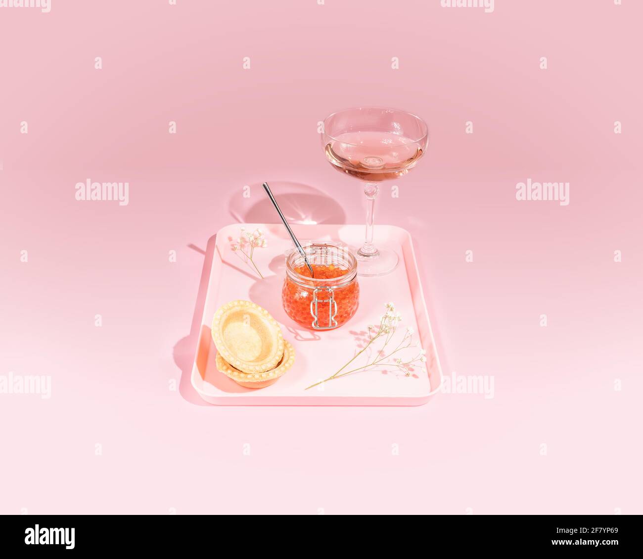 Romantische Speisekomposition mit einem Glas lachsroten Kaviar, Törtchen, Rosenwein und Blumen auf einem Tablett auf einem rosa Hintergrund mit harten Schatten Stockfoto