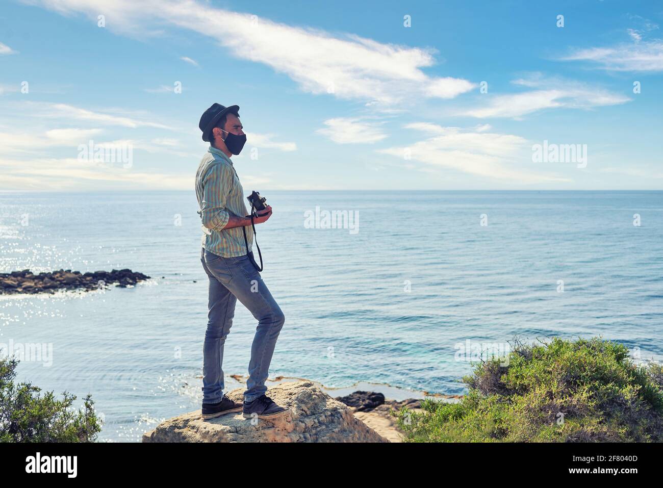 Ein junger Mann, Reisender und Abenteurer, hält seine Vintage-Kamera, trägt einen Hut und hat einen sehr coolen Hemd- und Jeans-Look, während er auf den Horizont blickt Stockfoto