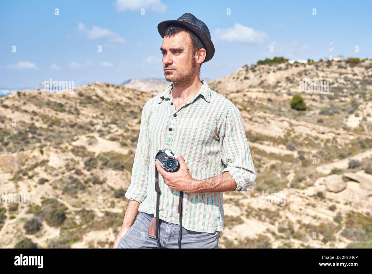 Ein junger Mann mit einem Hut des Reisenden und einem sehr Cool Look hält seine Kamera mit Bergen im Hintergrund Stockfoto