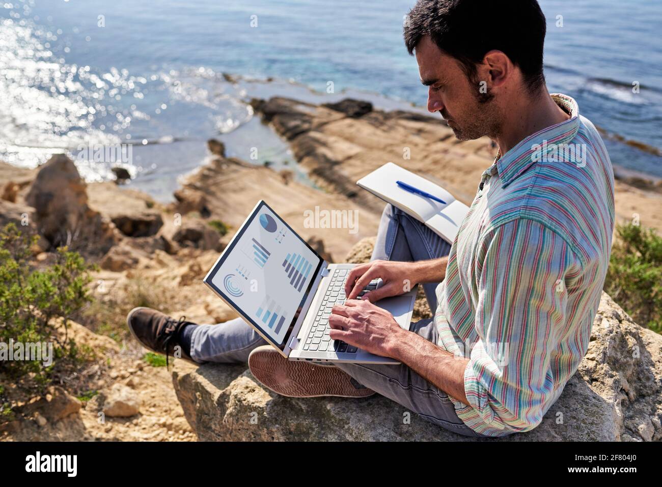Ein junger Mann benutzt während der Arbeit seinen Laptop und Notizblock Entfernt mit dem Meer im Hintergrund Stockfoto