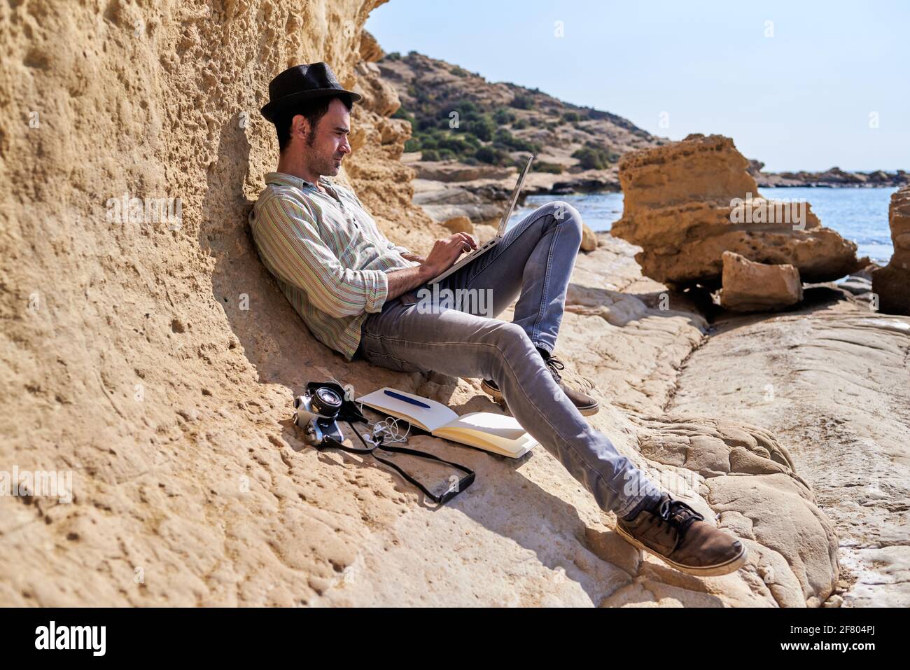 Ein junger Journalist entspannt sich beim Telearbeit vom Strand, neben ihm hat er eine Kamera, einen Notizblock und ein Smartphone. Stockfoto