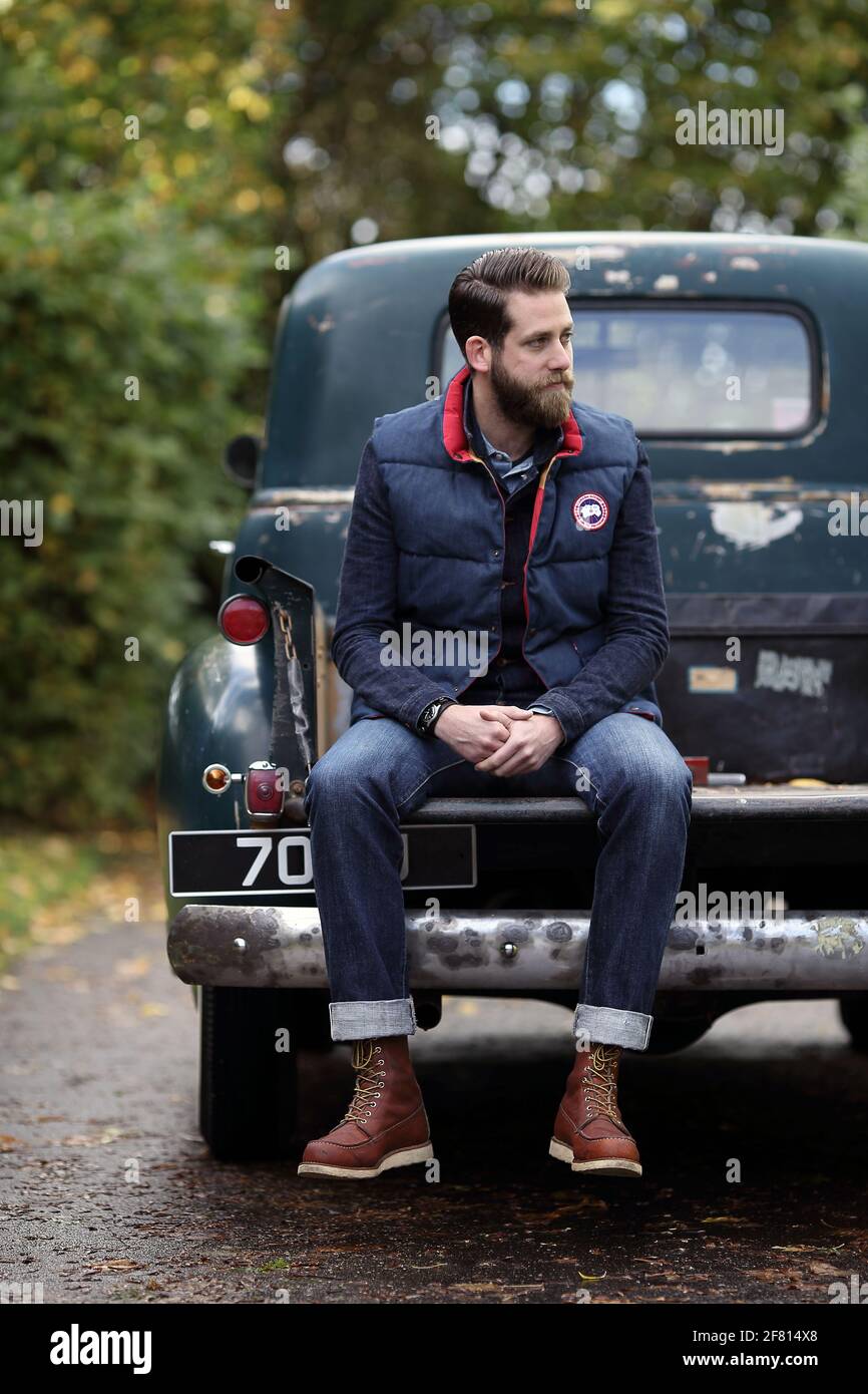 Berded junger Mann in Jeans sitzt auf seinem Pickup chevrolet Oldtimer draußen .junger Mann sitzt auf der Rückseite eines alten Chevrolet Pick-up-Truck. Stockfoto