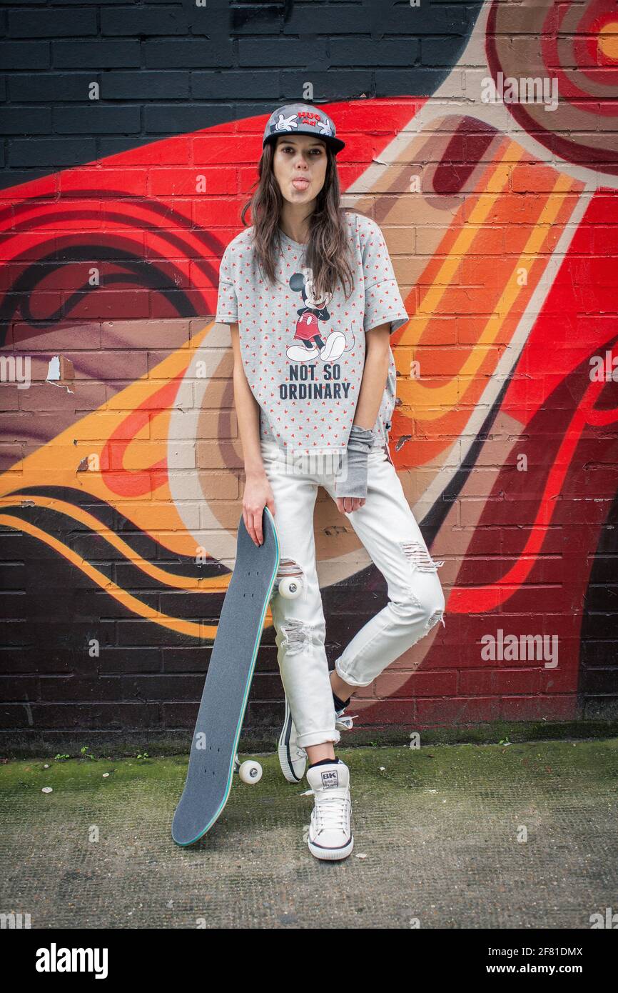 Teenage-Mädchen mit Skateboard, die Zunge herausstreckt und in der Nähe einer Stadtmauer mit bunten Graffiti in Shorditch in London, Großbritannien, steht. Stockfoto