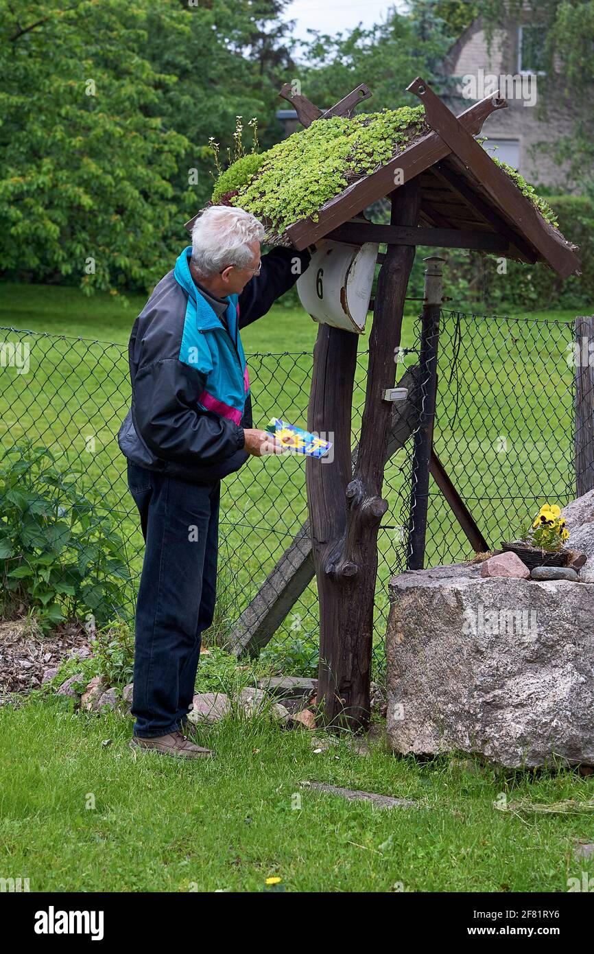 Seitenansicht eines älteren Mannes, der eine christliche Broschüre in einen deutschen Briefkasten legte, der mit einer moosigen überdachten Konstruktion in der Nähe eines Kettengliederzauns bedeckt war. Stockfoto