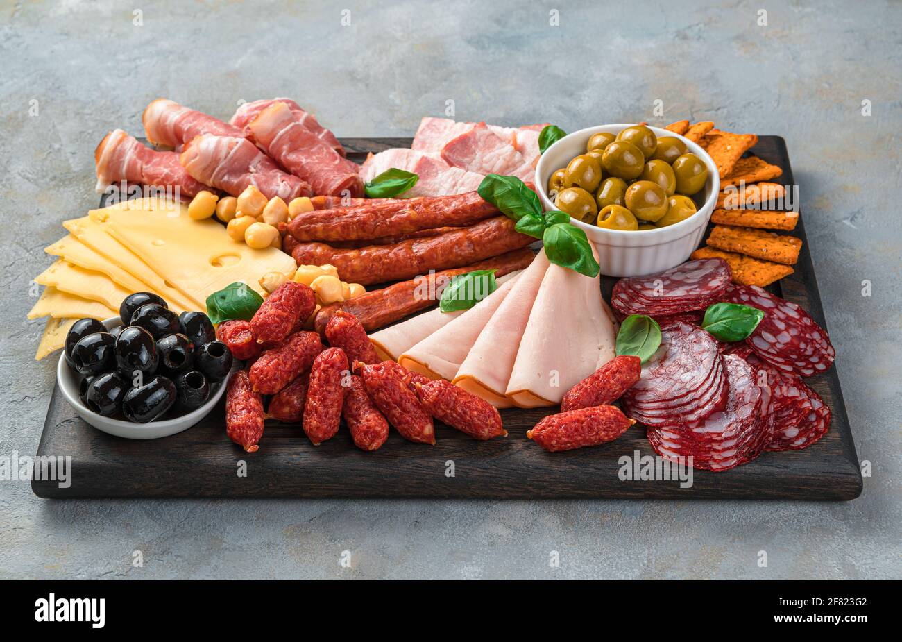 Vorspeisen-Bretter, Fleisch- und Käseschneiden mit Basilikum und Oliven auf einem rechteckigen Brett auf grauem Hintergrund. Seitenansicht. Stockfoto