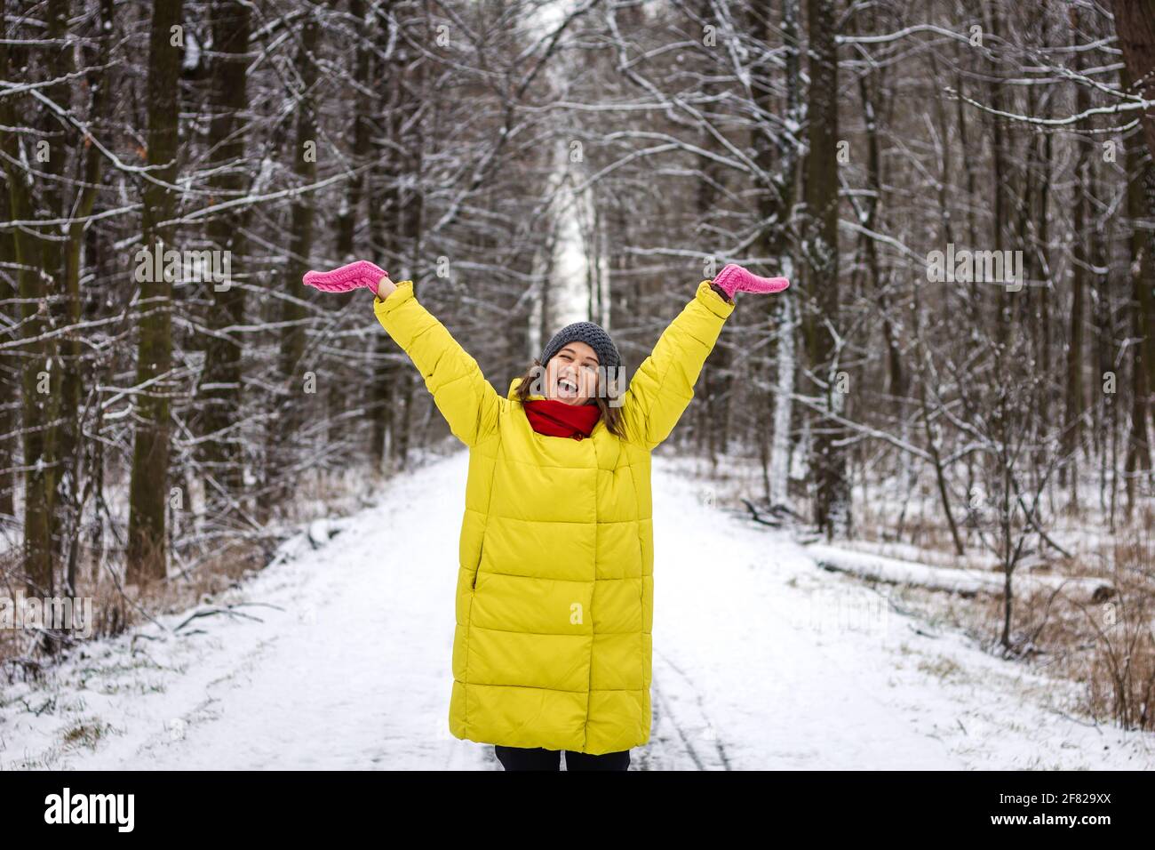 Glückliche junge Frau genießt Winterspaziergang im Wald. Frau in gelbem Mantel und Strickmütze in verschneite Natur Stockfoto