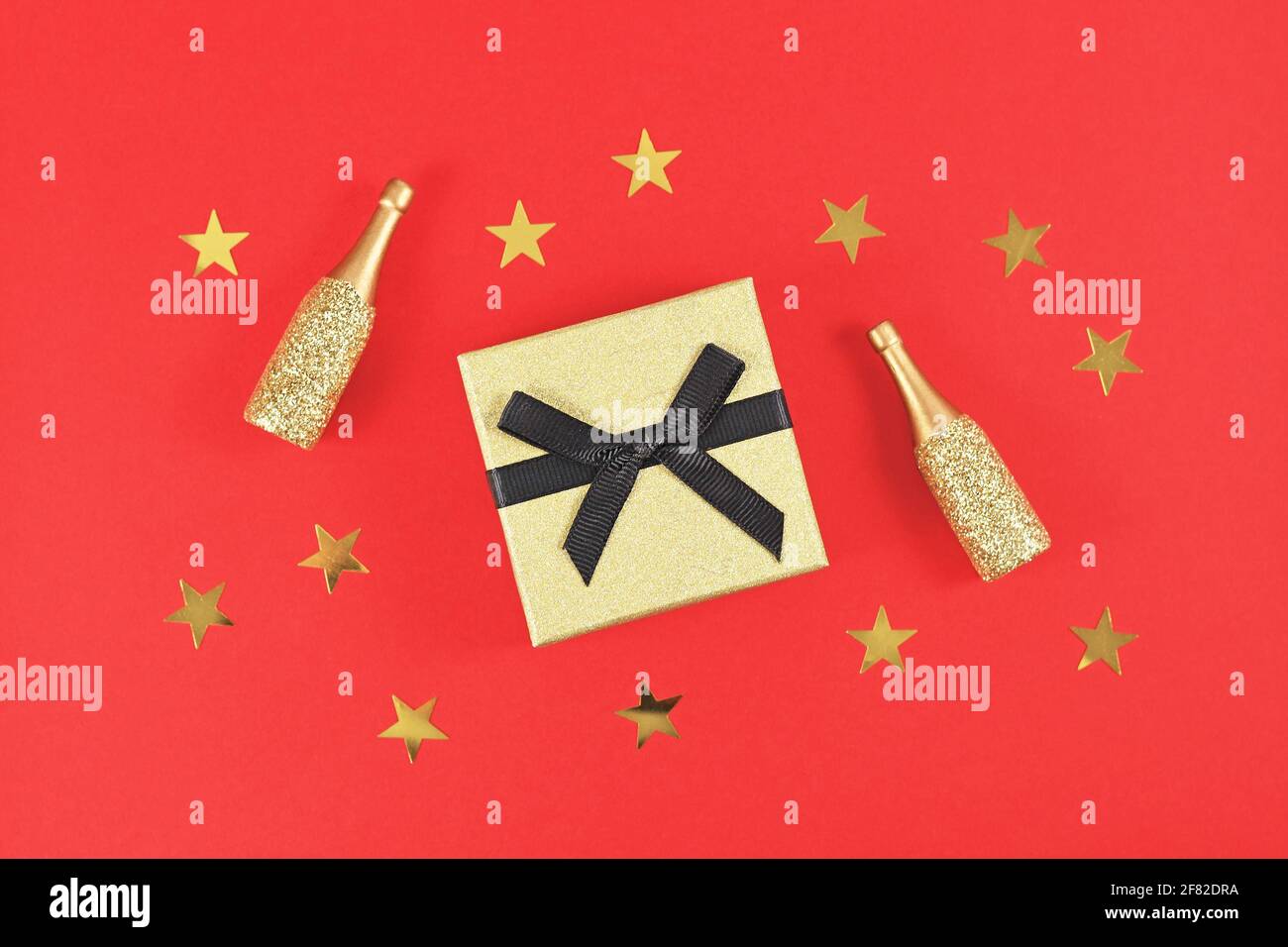 Goldene Geschenkbox mit schwarzer Schleife, kleinen Champagnerflaschen und Sternen auf rotem Hintergrund Stockfoto