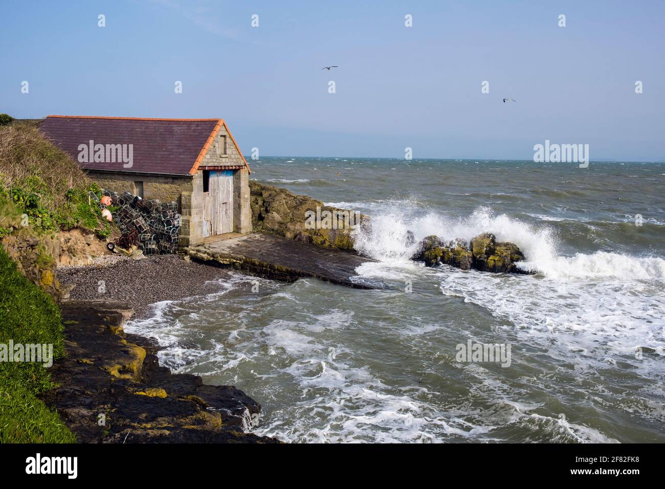 Raues Meer, das bei Flut am Bootshaus der alten Rettungsbootstation an einem windigen Tag auf Felsen stürzt. Moelfre, Isle of Anglesey, Nordwales, Großbritannien Stockfoto