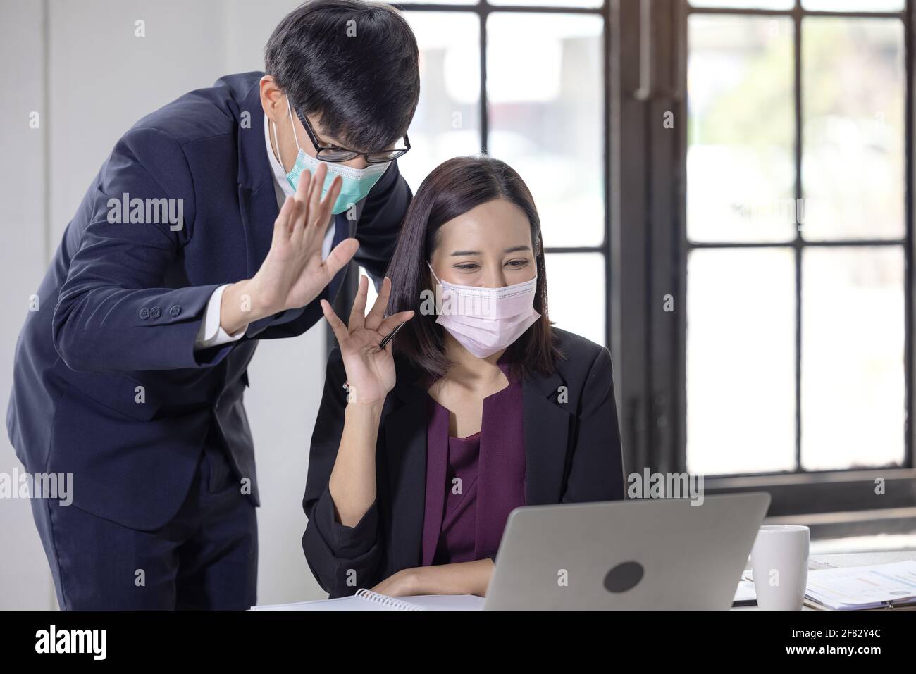 Geschäftsmann und Frauen tragen Gesichtsmasken, während sie ihren Laptop benutzen und jemanden während eines Videoanrufs im Büro begrüßen. Konferenz mit dem Arbeitsteam inmitten von COVID Stockfoto