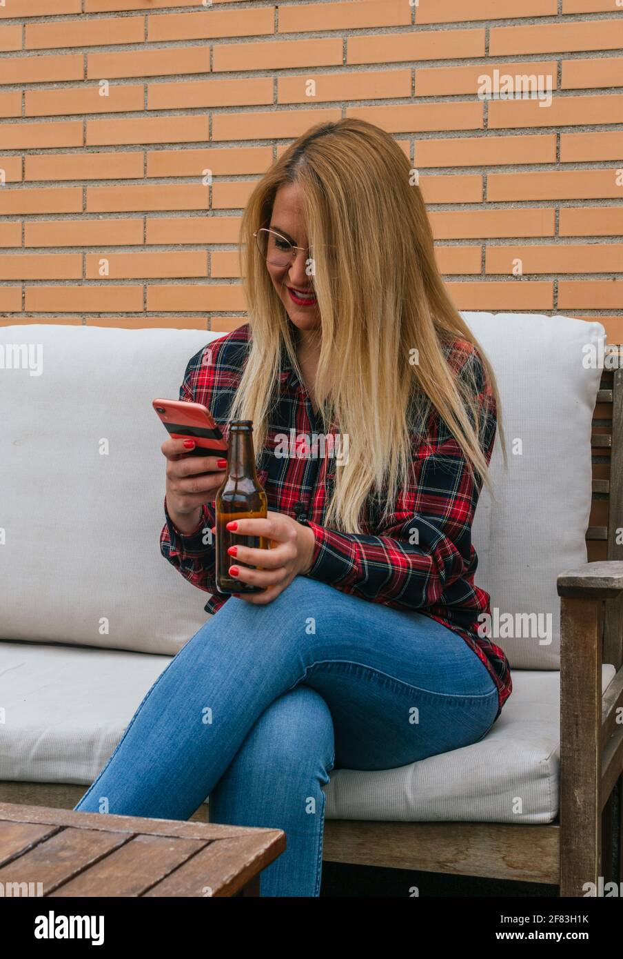 Junges blondes Mädchen, das mobil zusieht, lächelt und ein Bier auf der Terrasse trinkt Stockfoto