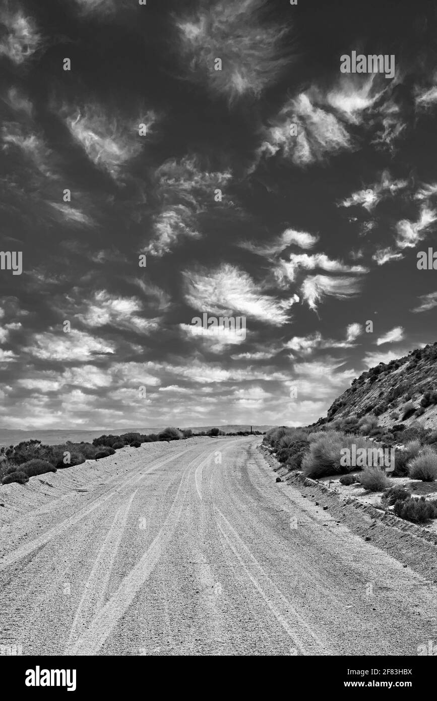 Einsame Wüste Schotterstraße in Richtung Horizont neben Hügeln. Weiße, flauschige Wolken am Himmel. Schwarz und Weiß. Stockfoto