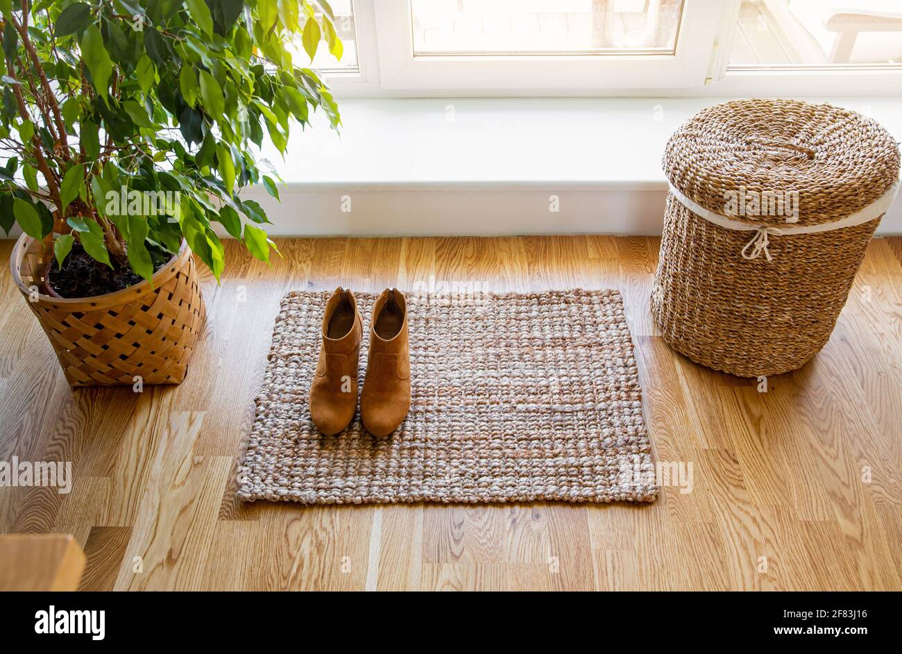 Hartholzboden mit Jute-Fußmatte, Schuhen und Blumentopf und Seegraswaschkorb am Fenster. Natürliche materielle Objekte im Heimkonzept. Inneneinrichtung. Stockfoto