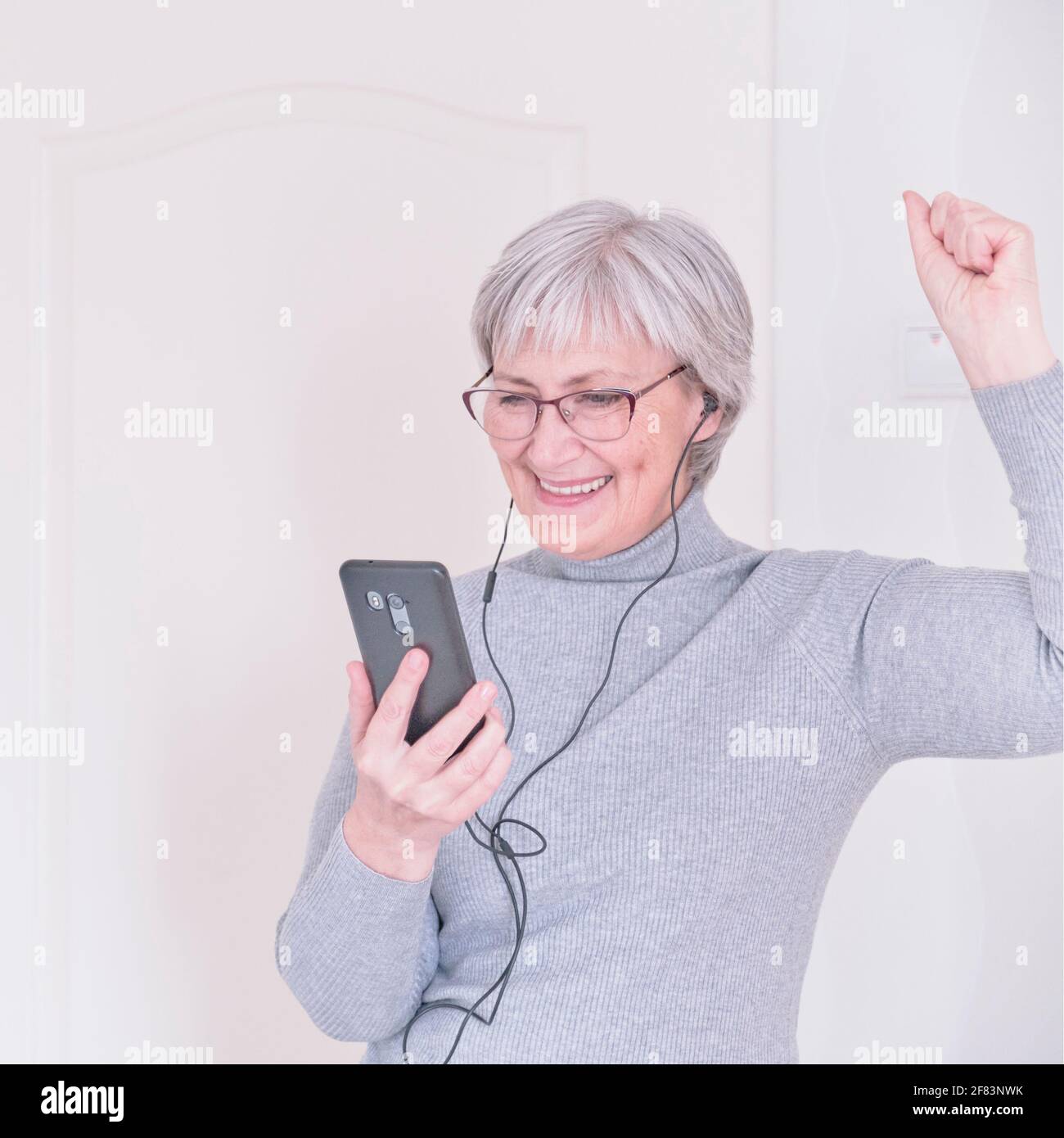 Eine grauhaarige ältere Frau mit Brille, in Kopfhörern, in einem grauen Rollkragen, die zu Hause auf das Smartphone schaut und Freude hat. Lifestyle-Moment. Stockfoto