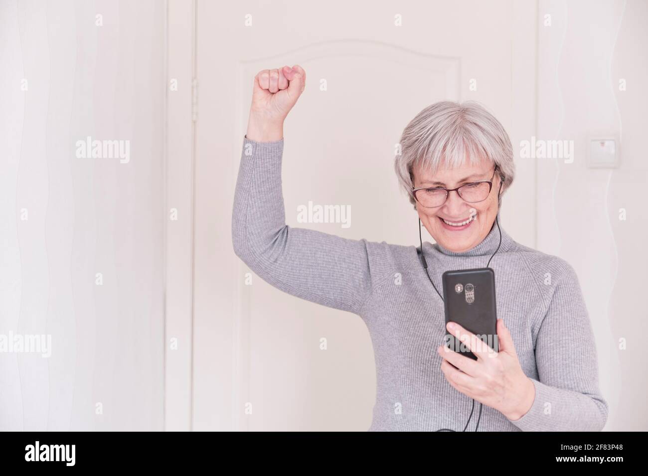 Eine grauhaarige ältere Frau mit Brille, in Kopfhörern, in einem grauen Rollkragen, die zu Hause auf das Smartphone schaut und Freude hat. Lifestyle-Moment. Stockfoto