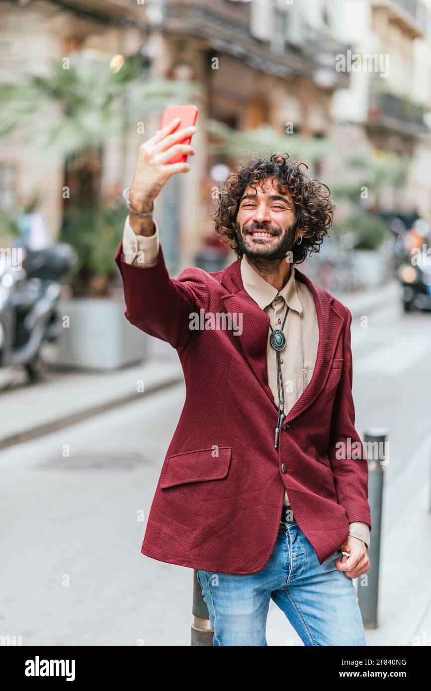 Der junge Kaukasusmann porträtiert sich mit seinem Telefon in den Straßen barcelonas. Er lächelt. Er hat lockiges Haar, Bart und trägt eine trendige rote Jacke und Jeans. S Stockfoto