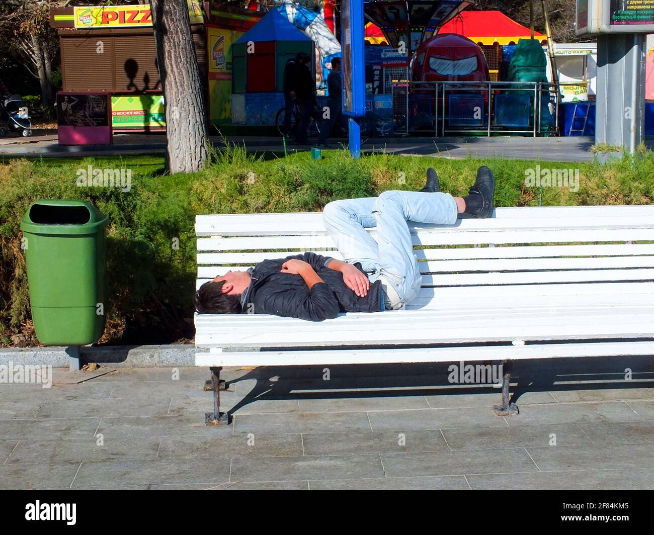 UKRAINE, JALTA - 29. JULI 2015: Obdachloser in Jeans und schwarzer Jacke schläft auf einer weißen Bank in der Nähe des grünen Mülltonnen auf der Straße. Stockfoto