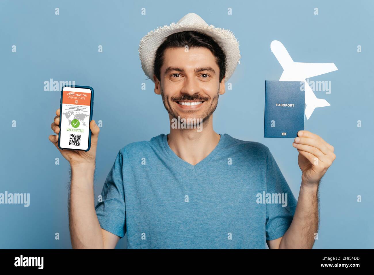 Elektronisches Impfzertifikat für Coronavirus-Infektion COVID19. Glücklicher Kerl in einem Hut, hält einen Pass mit kleinem Flugzeug, und Smartphone mit elektronischem Zertifikat, blau isolierten Hintergrund Stockfoto