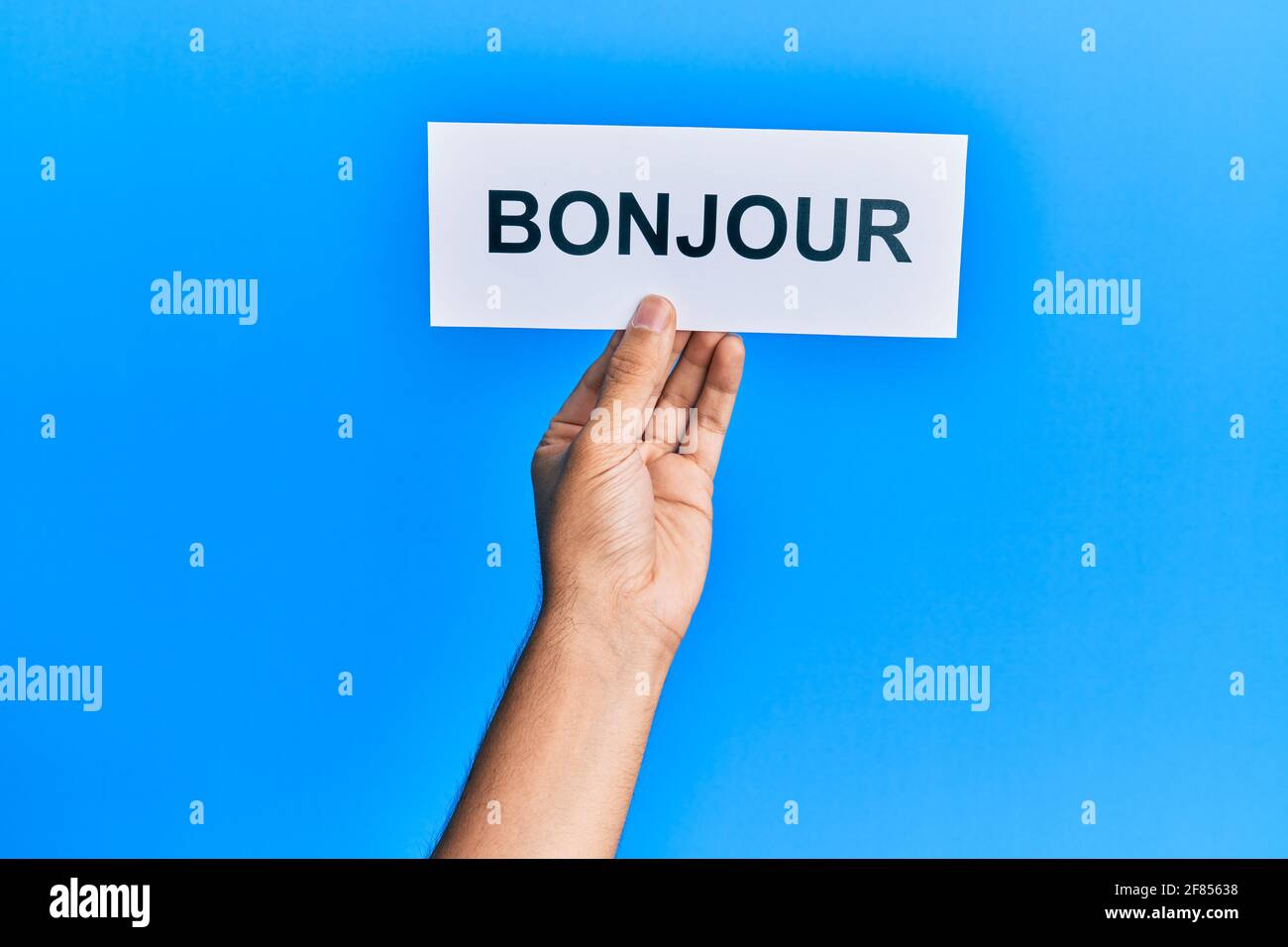 Hand von hispanischen Mann hält französisch Gruß bonjour Wort Papier über isolierten blauen Hintergrund. Stockfoto