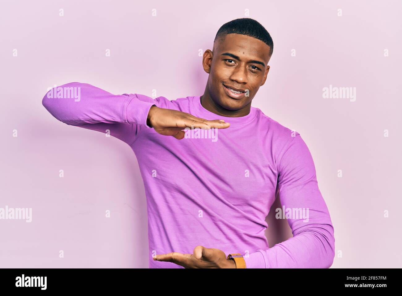 Junger schwarzer Mann trägt lässigen rosa Pullover gestikuliert mit Händen zeigt große und große Zeichen, Maßnahme Symbol. Lächelnd Blick auf die Kamera. Measu Stockfoto