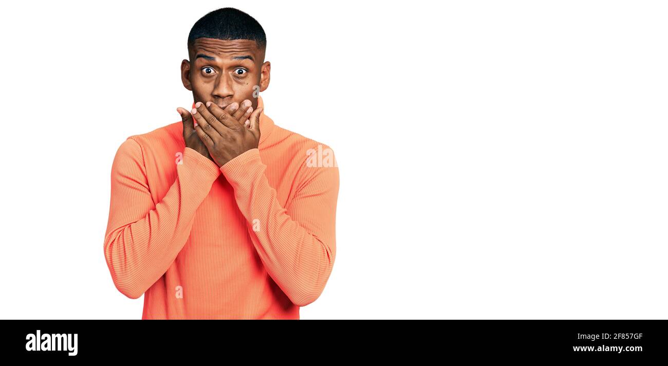 Junger schwarzer Mann mit orangefarbenem Rollkragenpullover schockiert, den Mund fälschlicherweise mit Händen zu bedecken. Geheimkonzept. Stockfoto