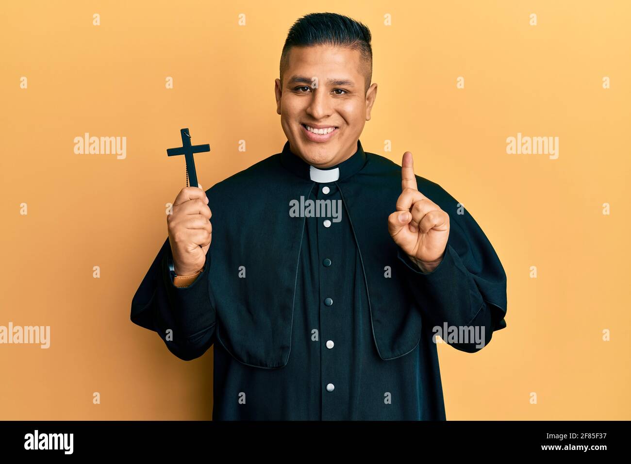 Junger lateinischer Priester, der Kruzifix hält, lächelt mit einer Idee oder Frage, die mit dem Finger und dem glücklichen Gesicht zeigt, Nummer eins Stockfoto