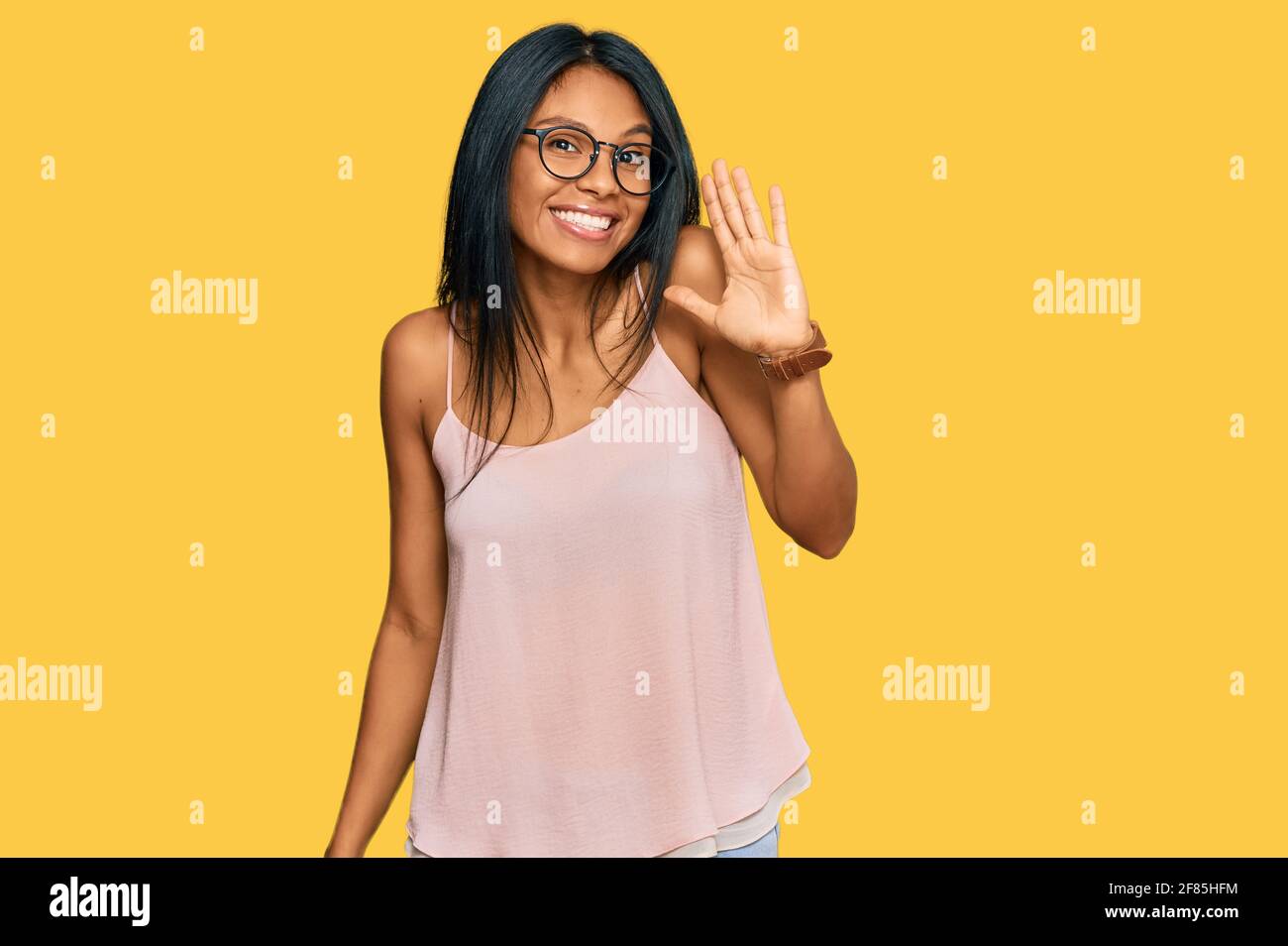 Junge afroamerikanische Frau trägt legere Kleidung und Brille waiving sagen Hallo glücklich und lächelnd, freundliche Willkommensgeste Stockfoto