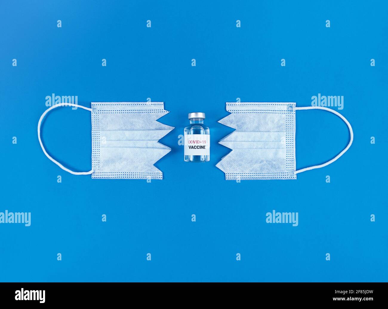 Fläschchen mit Covid19-Impfstoff zwischen zwei Teilen der geschnittenen medizinischen Maske auf blauem Hintergrund. Stockfoto