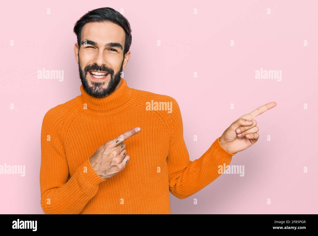 Junger hispanischer Mann in lässiger Kleidung lächelnd und mit zwei Händen und Fingern zur Seite auf die Kamera schauend. Stockfoto