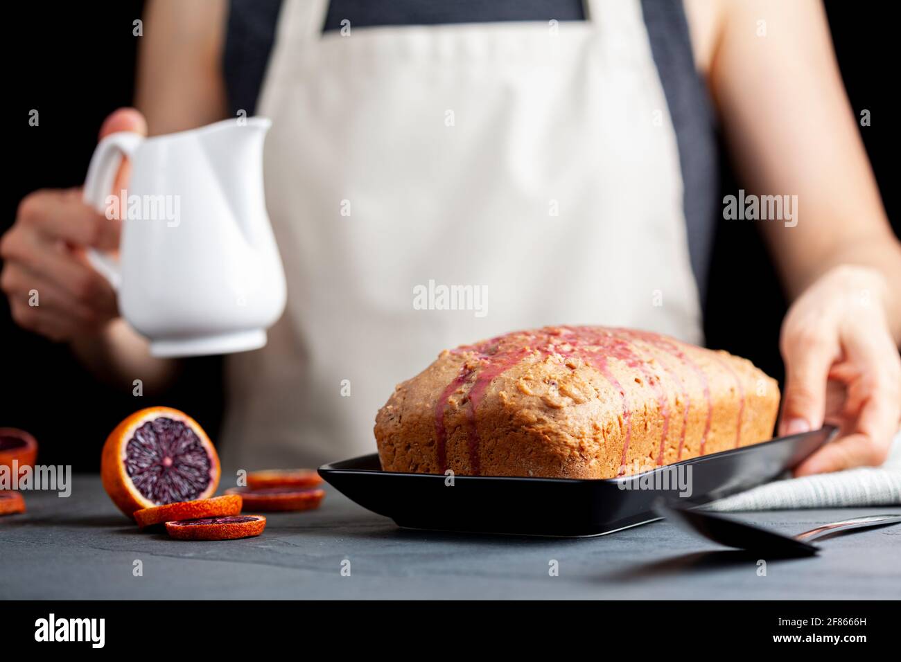 Nahaufnahme Bild von frischen hausgemachten Blut-Orange-Kuchen in einer Laib-Pfanne gemacht und serviert auf schwarzem Porzellan-Teller auf dunklem Stein Hintergrund. Eine Frau gießt Bloo aus Stockfoto