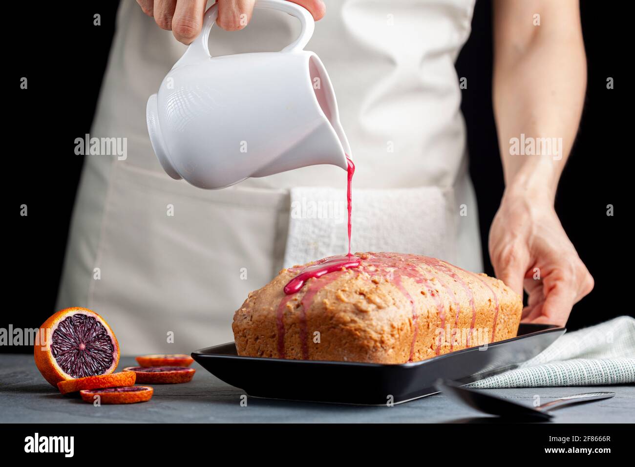 Nahaufnahme Bild von frischen hausgemachten Blut-Orange-Kuchen in einer Laib-Pfanne gemacht und serviert auf schwarzem Porzellan-Teller auf dunklem Stein Hintergrund. Eine Frau gießt Bloo aus Stockfoto