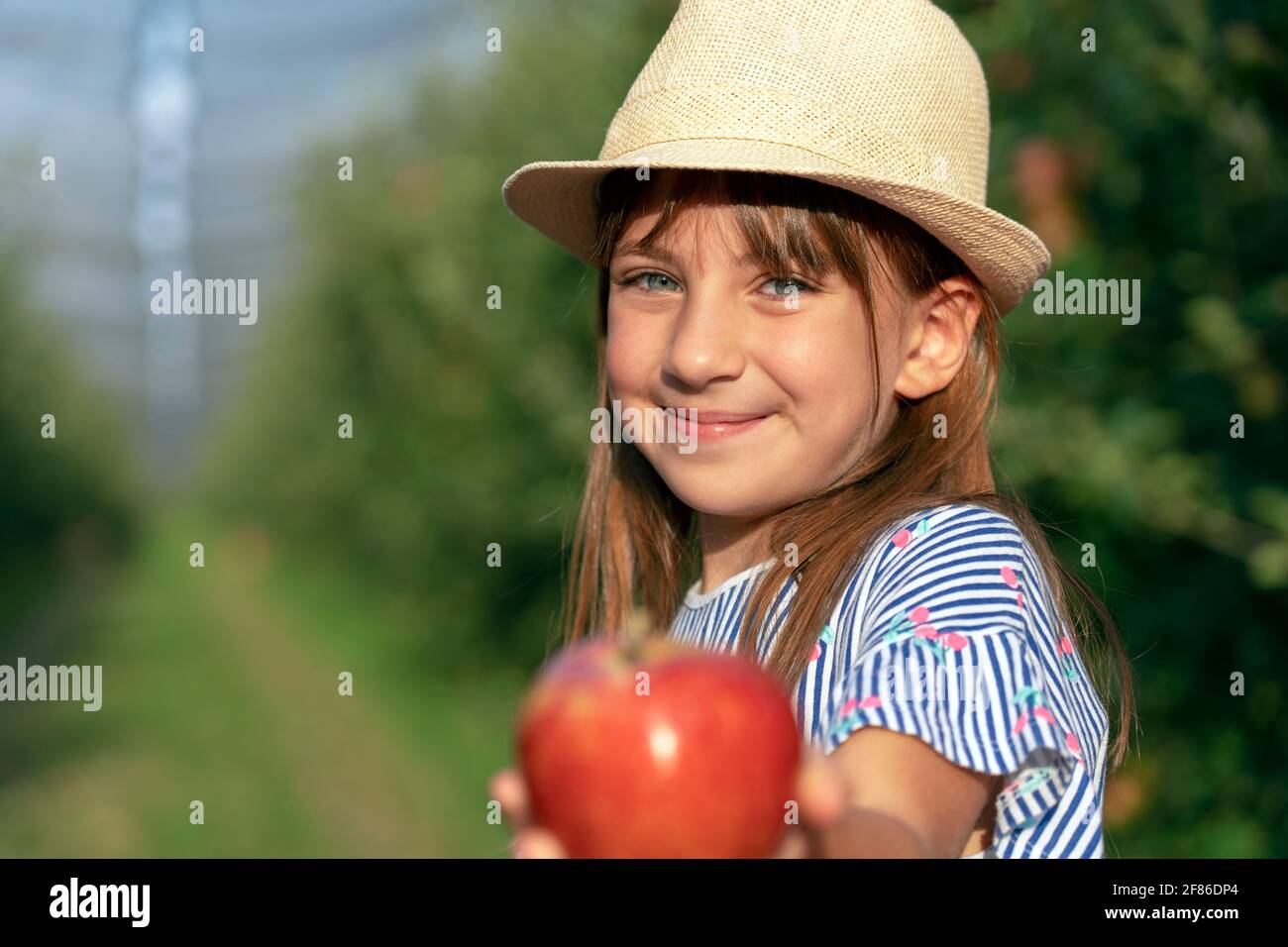 Porträt eines lächelnden kleinen Mädchens mit schönen Augen in einem Obstgarten. Konzept Für Gesunde Ernährung. Nettes Mädchen mit Hut hält Red Apple in der Hand. Stockfoto