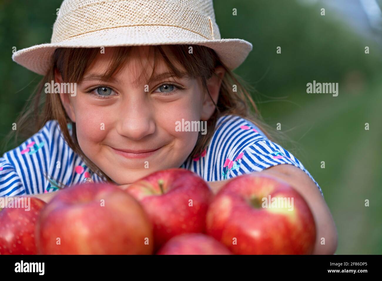 Lächelndes kleines Mädchen mit schönen Augen und Blick auf die Kamera. Konzept Für Gesunde Ernährung. Nahaufnahme des niedlichen Mädchens mit Hut und Korb mit roten Äpfeln. Stockfoto