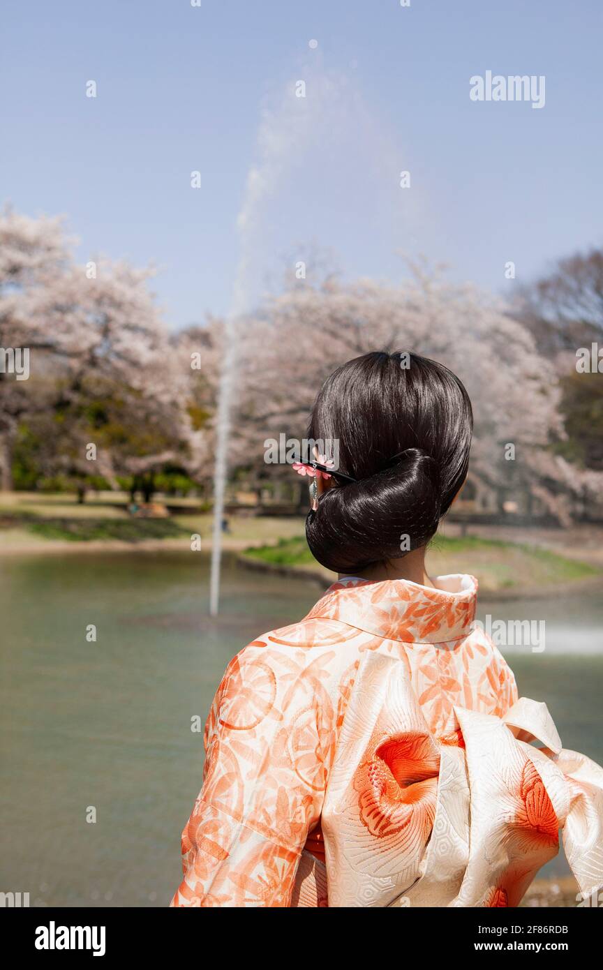 Junge Frau im orangefarbenen Kimono, die den Brunnen im sonnigen Frühling beobachtet parken Stockfoto