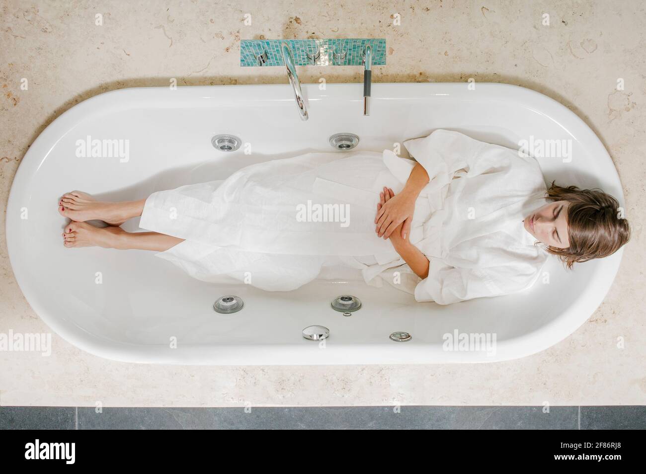 Ruhige junge Frau im Bademantel, die in der leeren Badewanne liegt Stockfoto
