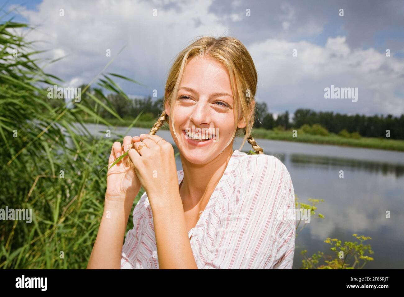 Portrait glückliche junge Frau mit blonden Zöpfen am sonnigen See Stockfoto