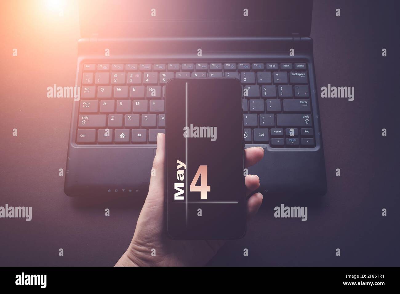 Mai. Tag 4 des Monats, Kalenderdatum. Hand halten Handy auf Laptop-Computer dunklen Hintergrund mit Sonnenschein. Frühling Monat, Tag des Jahres Konz Stockfoto