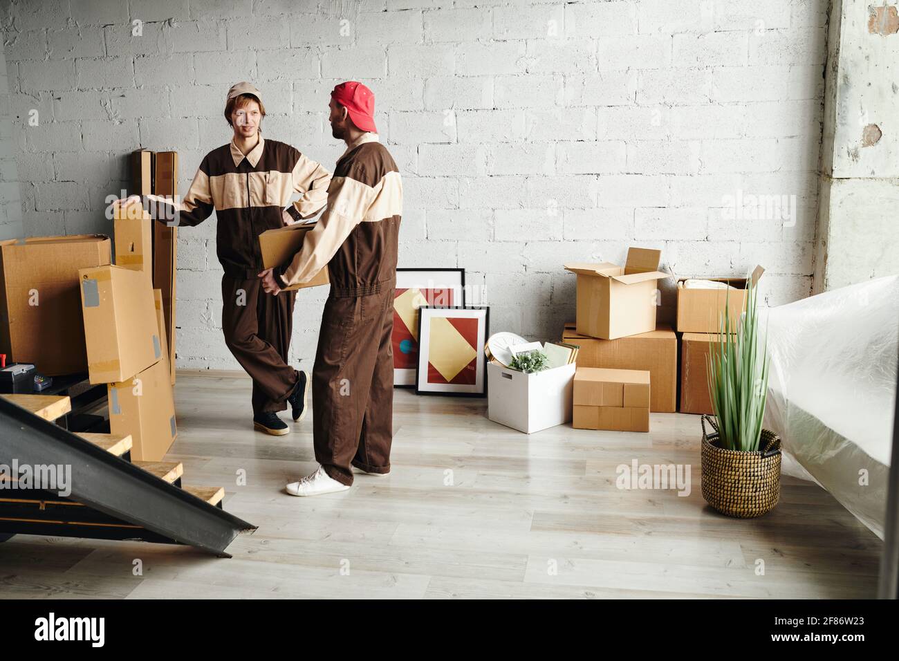Zwei junge Lader in Arbeitskleidung liefern verpackte Pappkartons und helfen Menschen beim Ausbauen in neue Wohnungen, Häuser oder Studios Stockfoto