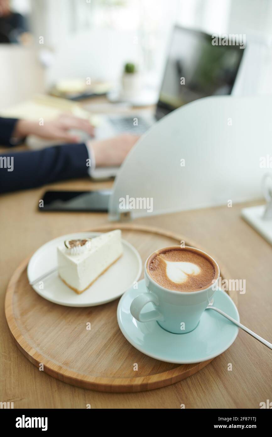 Tasse Cappuccino und Kuchensilice auf dem Kaffeetisch Von Geschäftsfrau, die am Laptop arbeitet Stockfoto