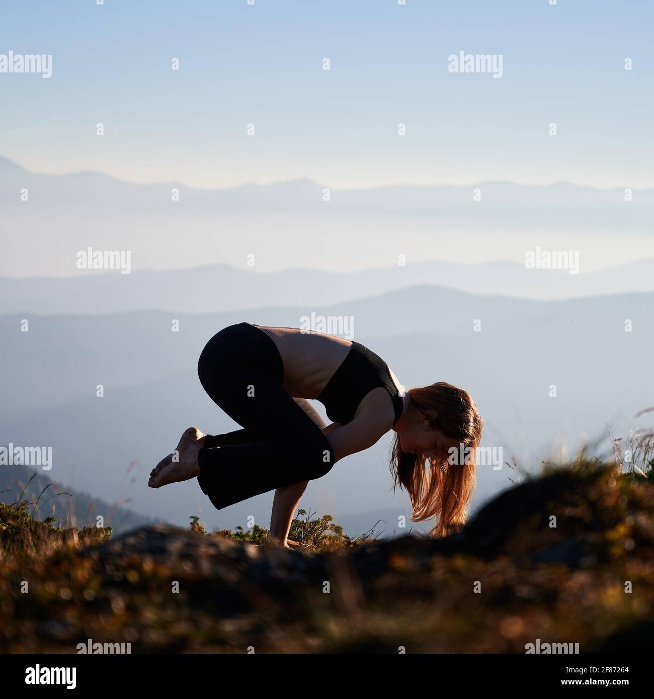 Sportliche junge Frau hebt Körper und balanciert auf den Händen, während Crow Pose auf grasbewachsenen Hügel am Morgen. Weibliche Person, die Yoga im Freien in Bergen mit blau-weißem Himmel auf dem Hintergrund praktiziert. Stockfoto