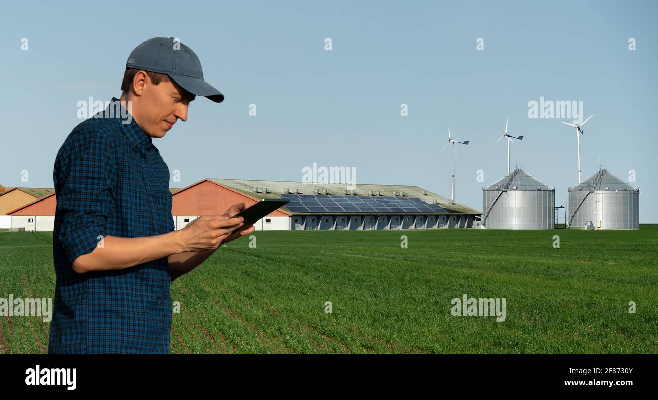Landwirt mit Tablet-Computer auf dem Hintergrund eines modernen Milchviehbetriebs mit erneuerbarer Energie, Solarzellen und Windturbinen Stockfoto