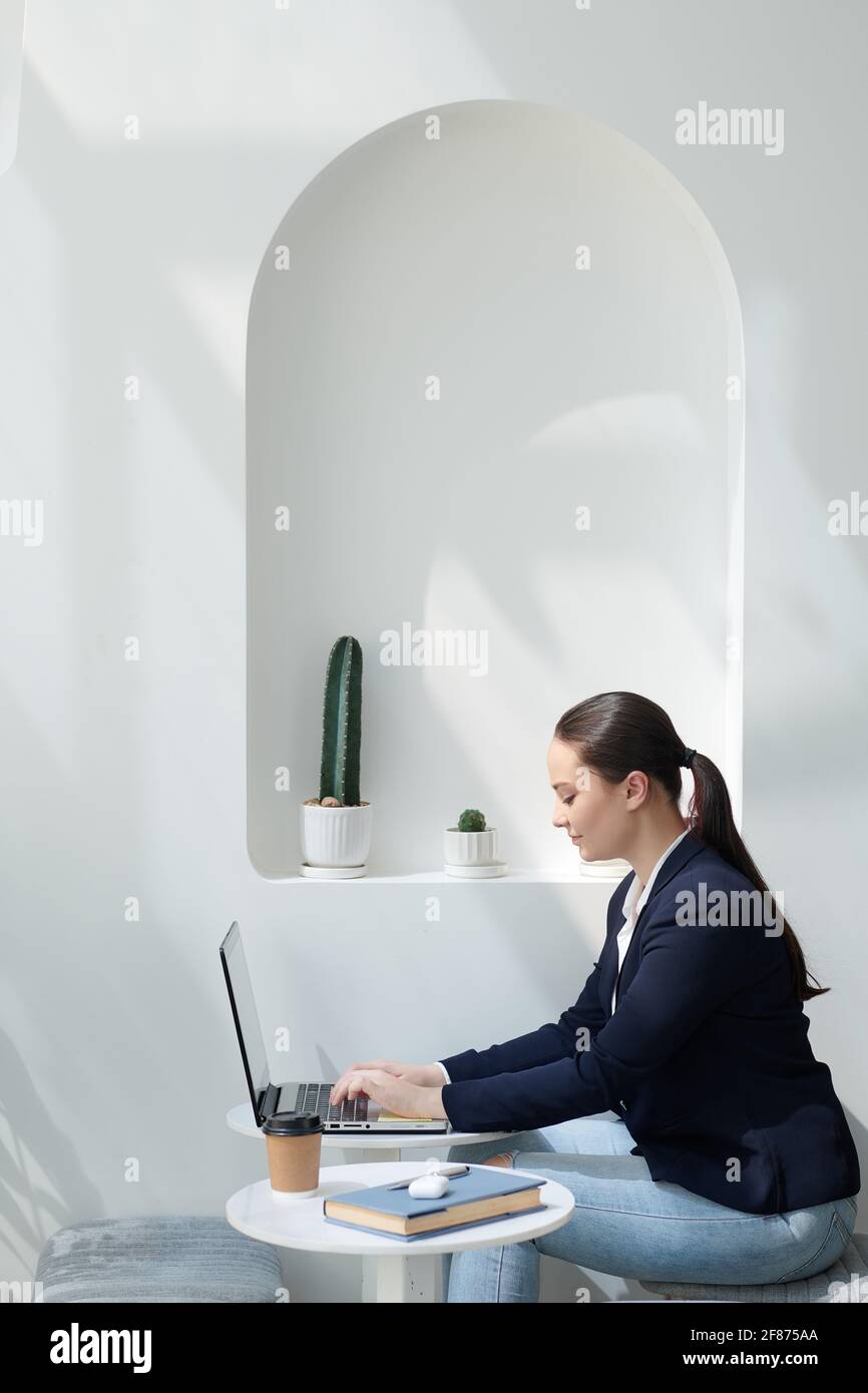 Lächelnde junge Unternehmerin, die in einem modernen Café am Tisch sitzt Und die Beantwortung von E-Mails auf dem Laptop Stockfoto