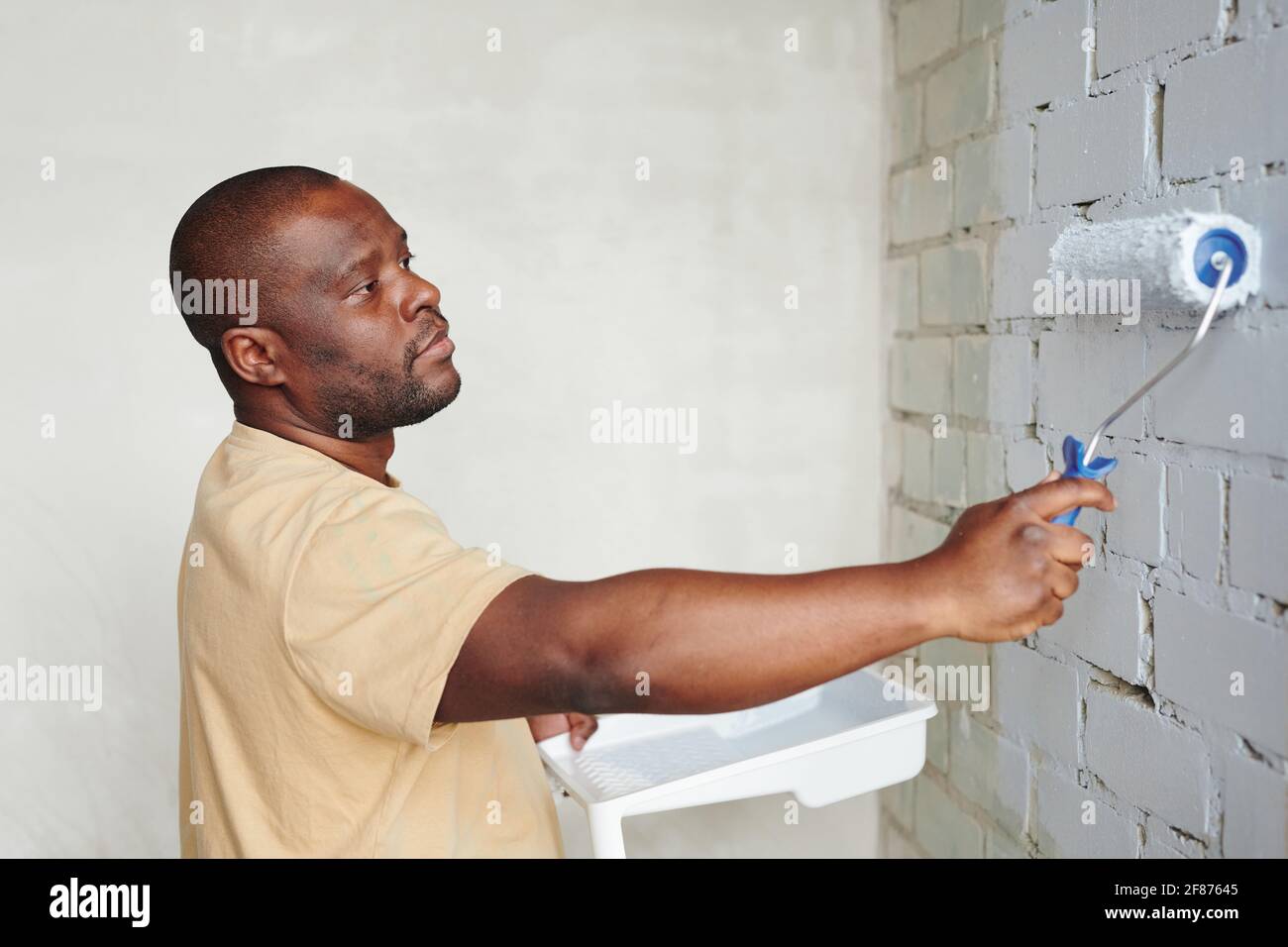 Junger afrikanischer Mann in beigem T-Shirt, der im Stehen den Paintroller hält Vor der Backsteinwand des Wohnzimmers und Bemalung In weiße Farbe Stockfoto