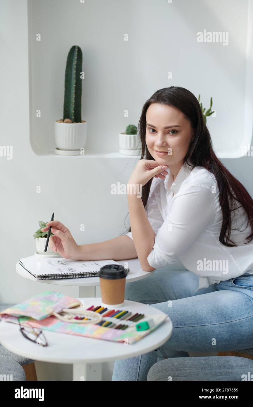 Ziemlich lächelnde junge Frau, die Zeit damit verbringt, Bilder in Skizzenbüchern zu zeichnen Mit Buntstiften und Tintenstiften Stockfoto