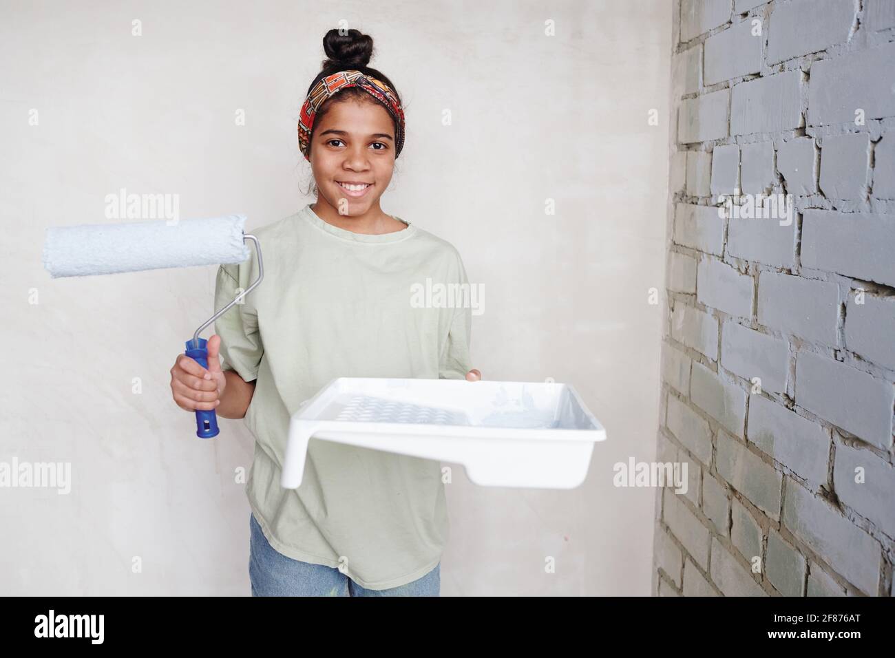 Happy niedlichen Teenager-Mädchen mit Paintroller und weißen quadratischen Kunststoff Behälter mit Farbe, die in der Ecke an zwei Wänden steht Des Wohnzimmers Stockfoto