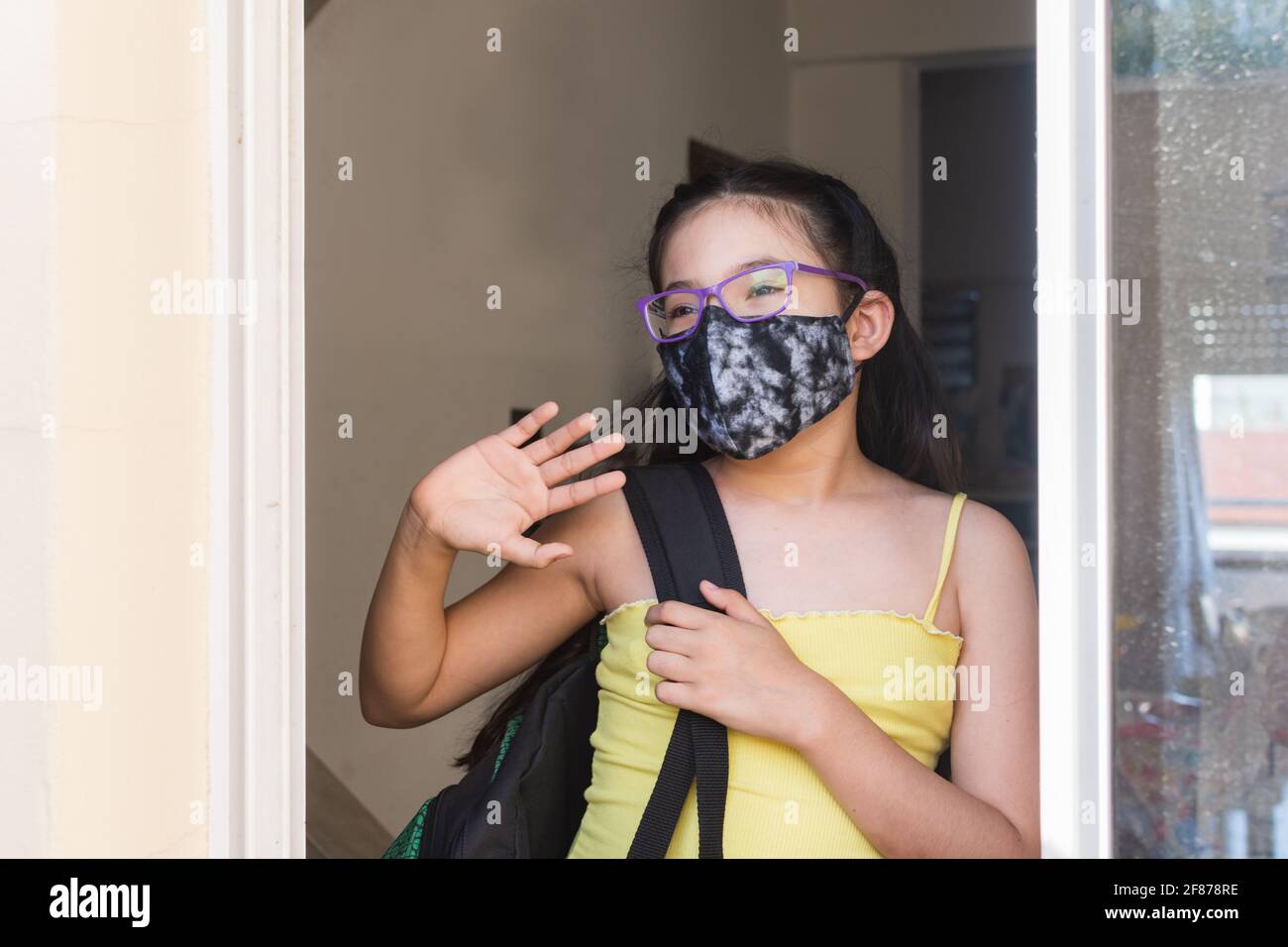 Vertikale Aufnahme eines kleinen hispanischen Mädchens mit einer Maske Und eine Brille, die jemandem Auf Wiedersehen sagt Stockfoto