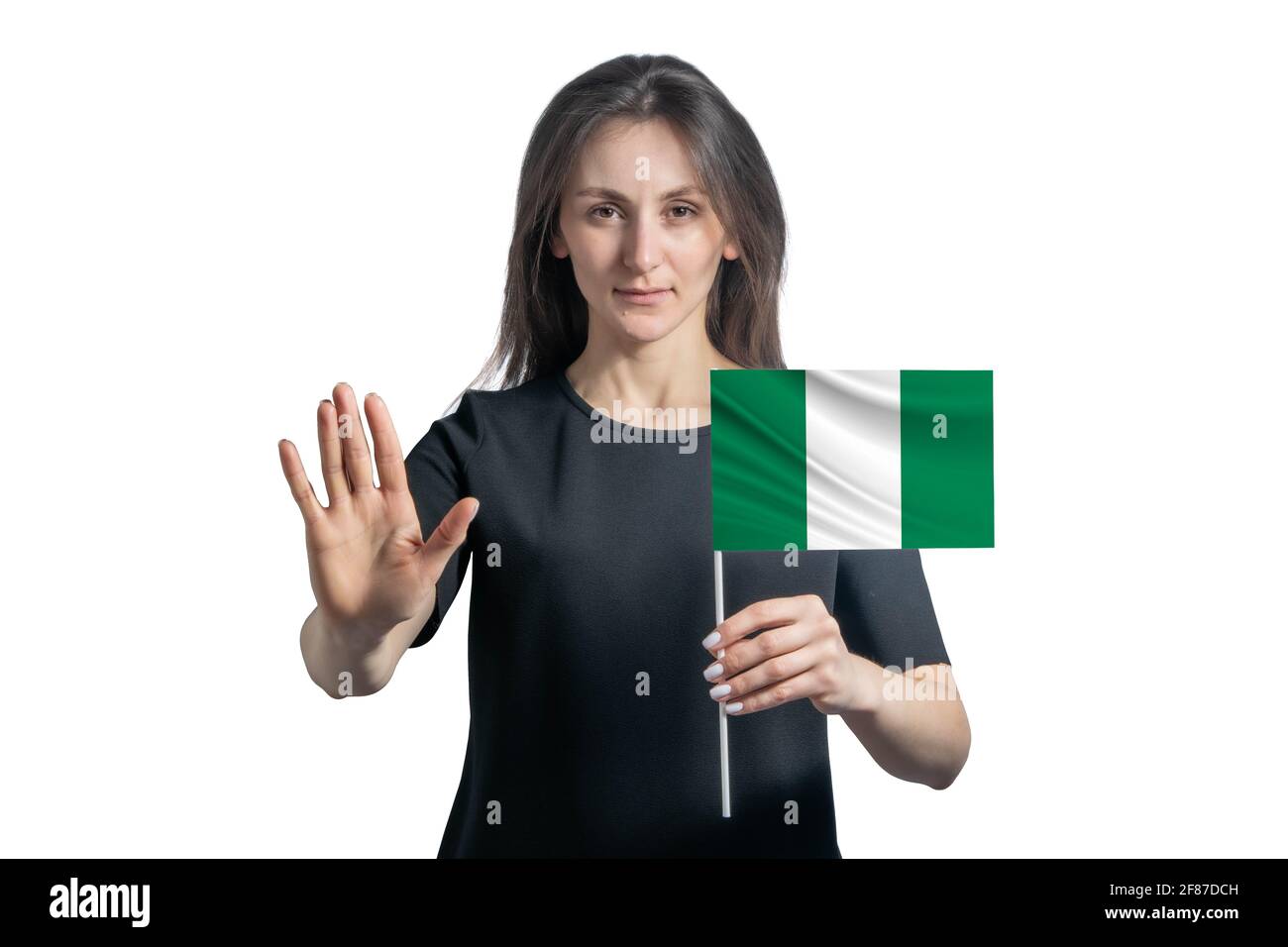 Glückliche junge weiße Frau, die die Flagge von Nigeria hält und mit einem ernsten Gesicht ein Hand-Stopp-Schild auf weißem Hintergrund zeigt. Stockfoto