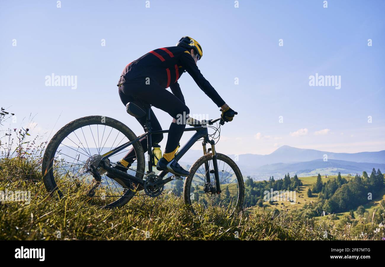 Horizontale Momentaufnahme eines Mannes, der am frühen nebligen Morgen mit seinem Fahrrad in den Bergen fährt und bergab fährt. Gipfel in Wolken, grüne Hügel im Hintergrund. Rückansicht, Kopierbereich. Konzept des aktiven Lebensstils Stockfoto